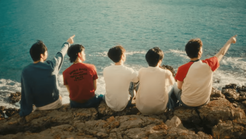Cena do MV de Love Language com os membros do TXT sentados de costas para a câmera, enquanto olham o mar