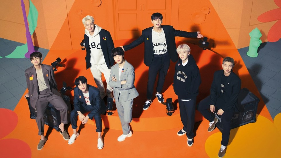 Imagem com todos os membros do BTS sorrindo em um cenário com o fundo laranja para o álbum Permission to Dance on Stage - Live