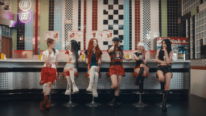 Cena do MV de "I WANt IT" com todas as integrantes do STAYC reunidas, lado a lado, em um ambiente colorido que lembra uma lanchonete americana