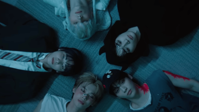 Cena do MV de "Beautiful Strangers" do álbum The Star Chapter: TOGETHER. Todos os membros do TXT estão deitados em um cenário escuro, câmera filmando de cima