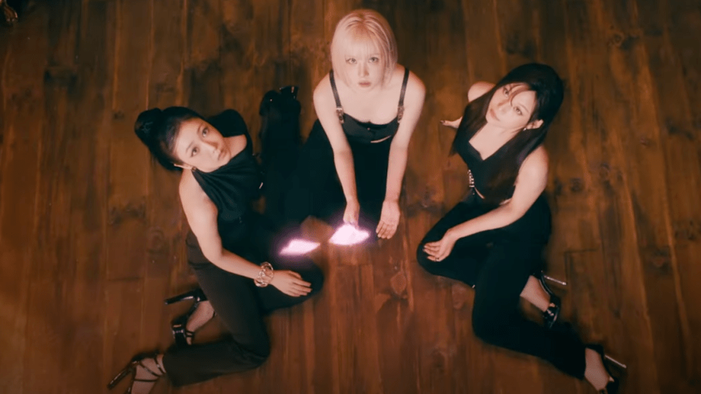 Cena do MV de "La La Love Me" do álbum A Montage of ( ) do VIVIZ com o trio sentado em um piso de madeira, enquanto olham uma borboleta brilhante voar