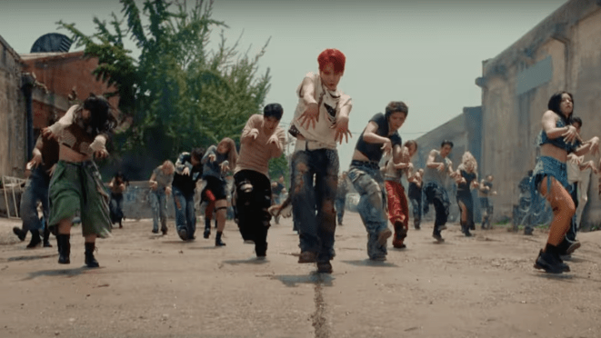 Cena do MV de "HUNTER" com Key de cabelo vermelho, dançando ao ar livre em um cenário caótico, com vários dançarinos ao seu redor