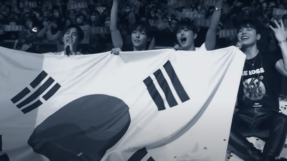 Cena do MV de "Trauma" do The Rose com os quatro segurando uma bandeira da Coreia do Sul