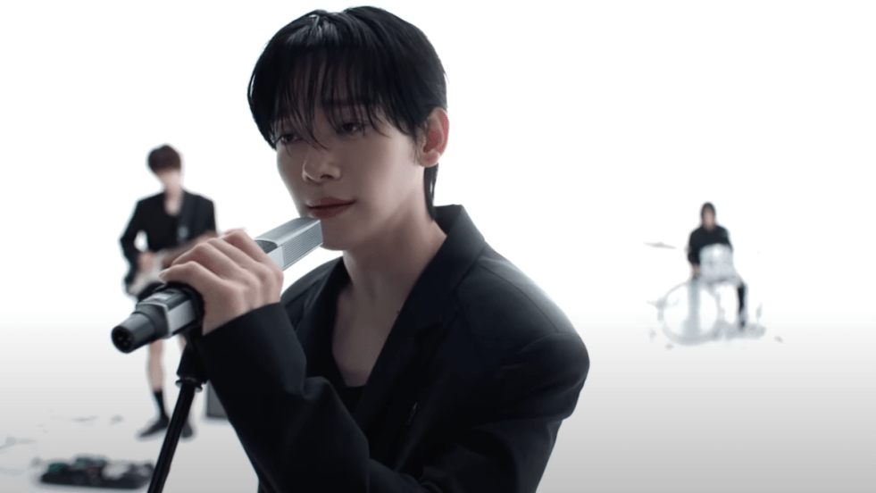 Cena do MV de "Arcadia" com Chen cantando enquanto segura um microfone. Roupas pretas e cenário todo branco