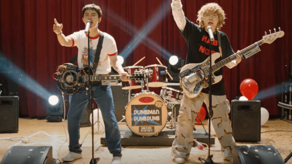 Cena do MV de "DUMB", single de D.O. do EXO e Penomeco, com os dois no palco e guitarras no pescoço