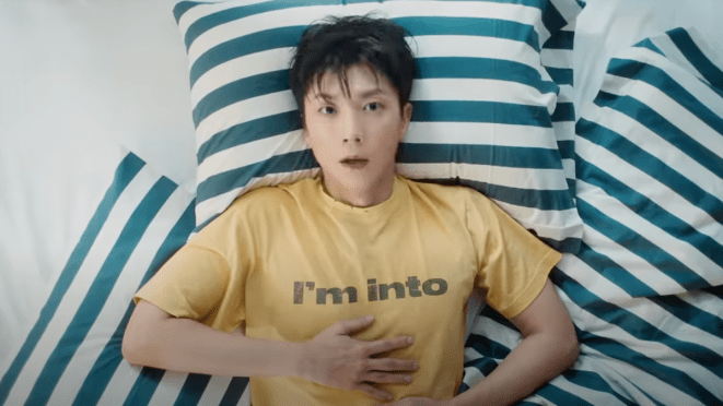Cena do MV de "Think Too Much" do álbum I'm Into do Jang Wooyoung [2PM]. Na imagem, ele aparece deitado em almofadas listradas com uma camiseta amarela que mostra o nome do álbum