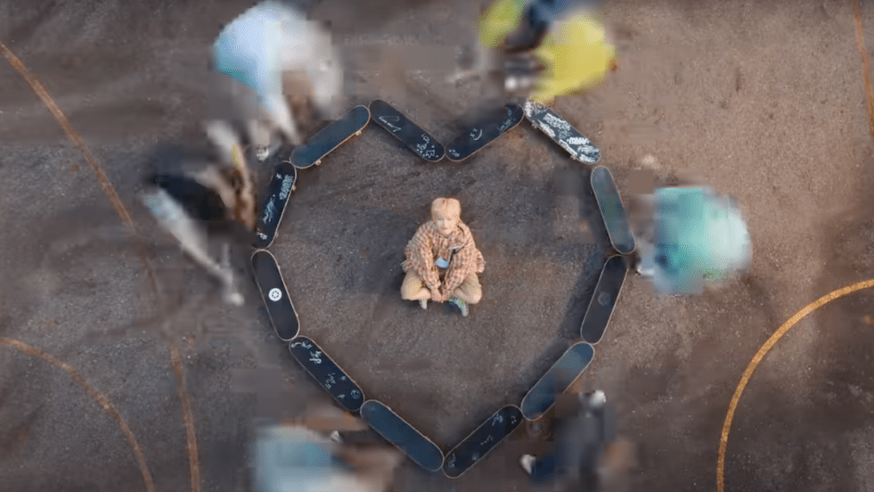 Cena do MV de “My First Love Song (처음 불러보는 노래)” do álbum My First Flip do KickFlip com um coração formado por skates e Keiju no centro