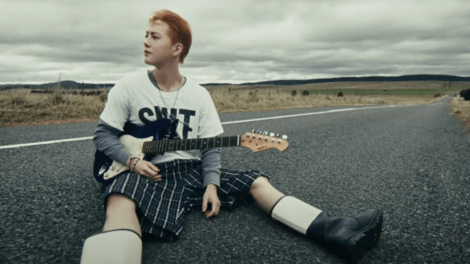 Cena inicial do MV de Who Are You do Suho [EXO sentado no meio da estrada, com uma guitarra no colo