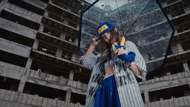 Cena do MV de "M.O." da Yuqi [i-dle]. Na imagem, ela segura uma sombrinha transparente em um cenário que parece um prédio abandonado