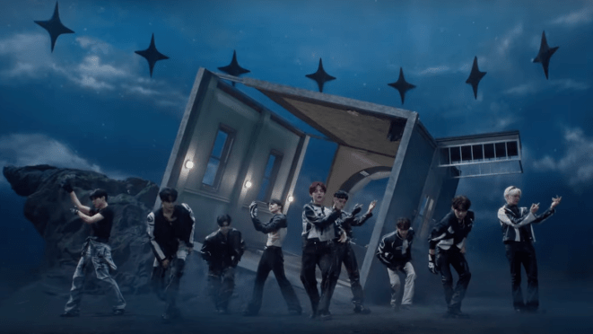 Cena do MV de "ICONIK" do álbum NEVER SAY NEVER do ZB1. Na imagem, todos dançam em um cenário noturno aberto com estrelas projetadas no céu