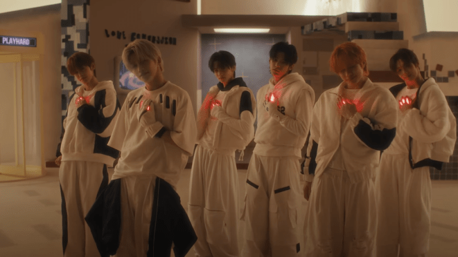 Cena do MV de "OVERDRIVE" do álbum play hard com todos os membros sorrindo, enquanto uma luz vermelha brilha no lugar do coração