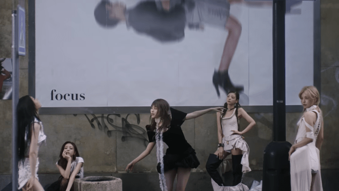Cena do MV de "TUNNEL VISION" do álbum homônimo do ITZY, com as cinco ao ar livre e um grande ao outdoor ao fundo com a palavra 'focus' em destaque