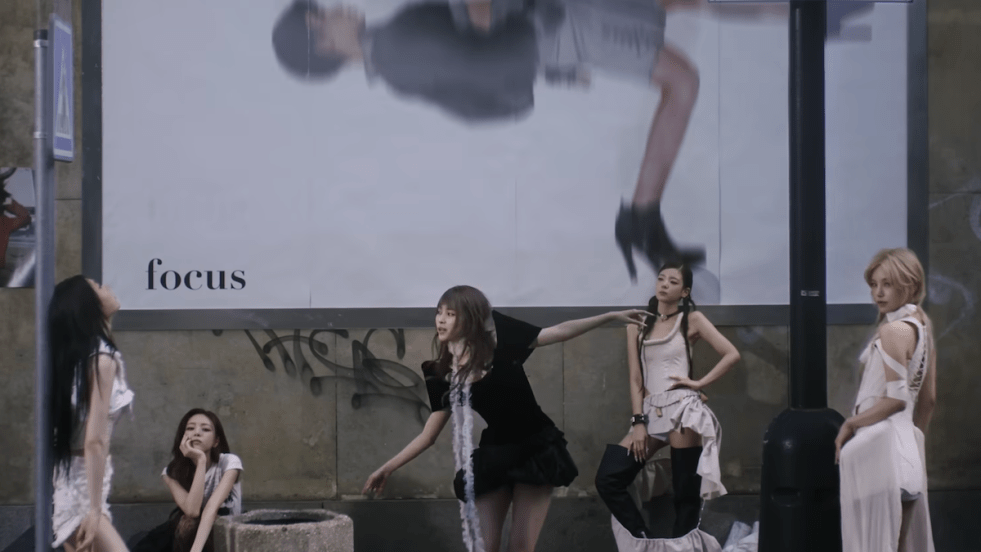Cena do MV de "TUNNEL VISION" do álbum homônimo do ITZY, com as cinco ao ar livre e um grande ao outdoor ao fundo com a palavra 'focus' em destaque