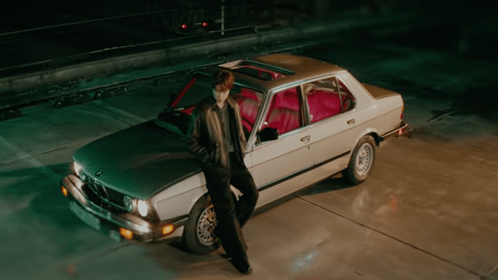 Cena do MV de "Backseat Promises" do álbum PULSEPHASE, com Kang Daneil encostado no capô de um carro durante a noite