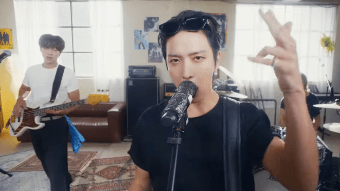 Review do álbum 3LOGY: cena do MV da title track "Killer Joy" com Jung Yong Hwa em foco, acompanhado por Kang Min Hyuk e Lee Jung Shin em um palco caseiro