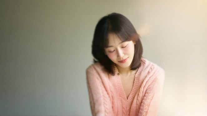 Review do K-Drama Love Me: Seo Hyun Jin (Seo Jun Kyung) com os olhos fechados e sorrindo em um cenário de cores quentes e com ar de aconchego