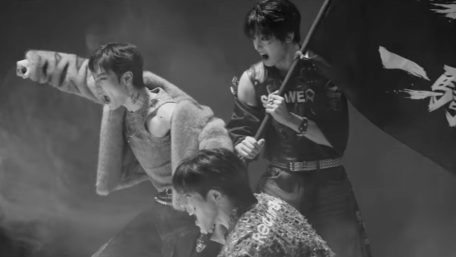 Review do álbum THE GREATEST BATTLE do Big Ocean: cena do MV com Chanyeon, Jiseok e PJ segurando uma bandeira e gritando como se estivessem rumo à vitória em uma guerra